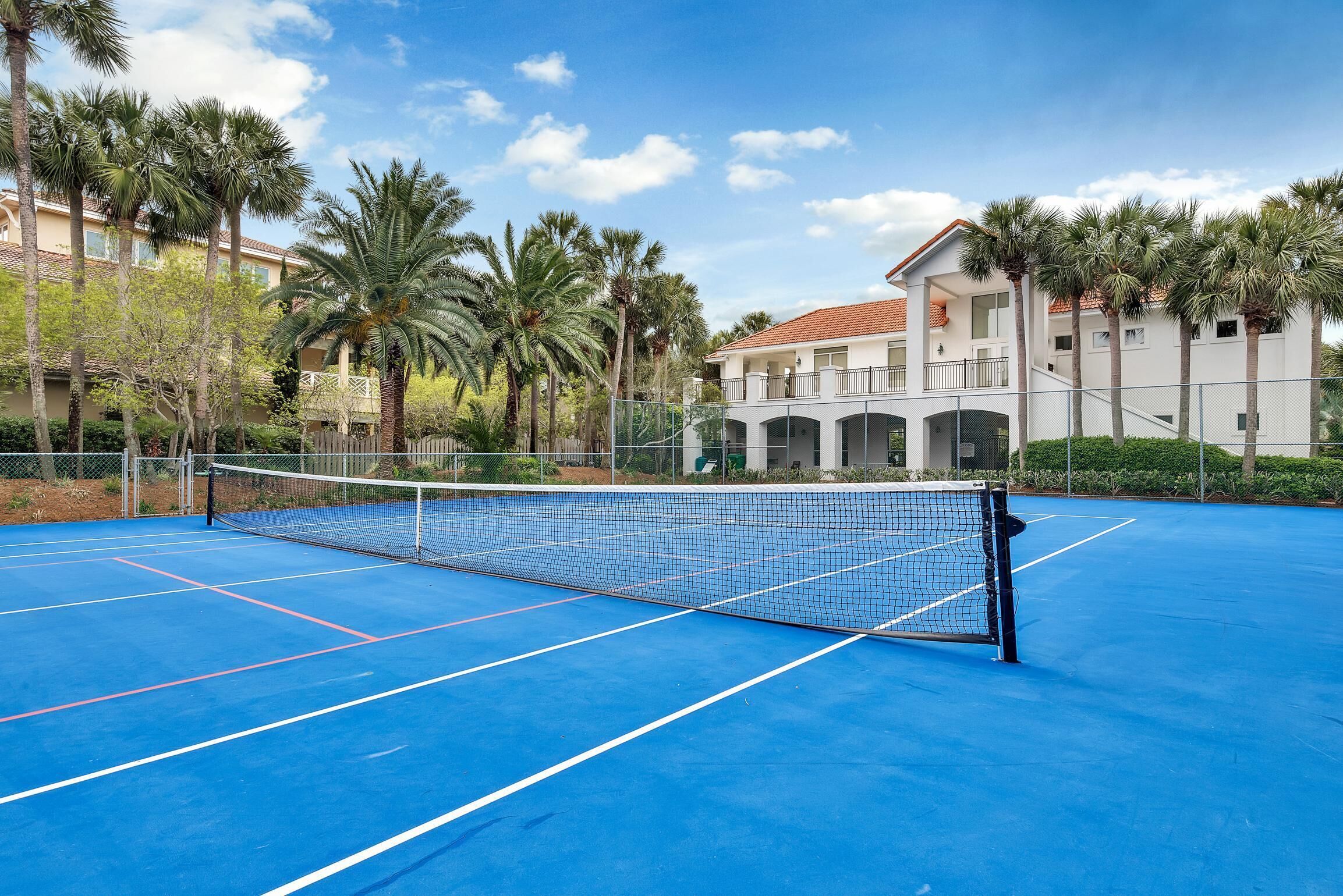 A tennis court with a net, surrounded by palm trees and a building with balconies.