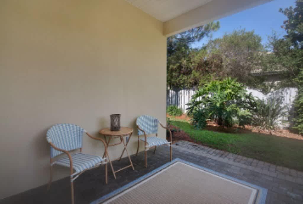 A patio with two wicker chairs and a small table, a rug, and a view of a garden with trees and a fence.