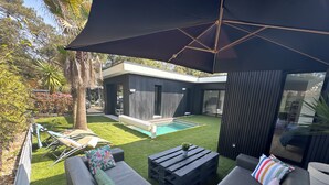Uma área de estar moderna ao ar livre com piscina, um espaço coberto para sentar e uma casa contemporânea ao fundo.