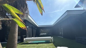 Uma área de estar moderna ao ar livre, com duas espreguiçadeiras, piscina e uma palmeira.