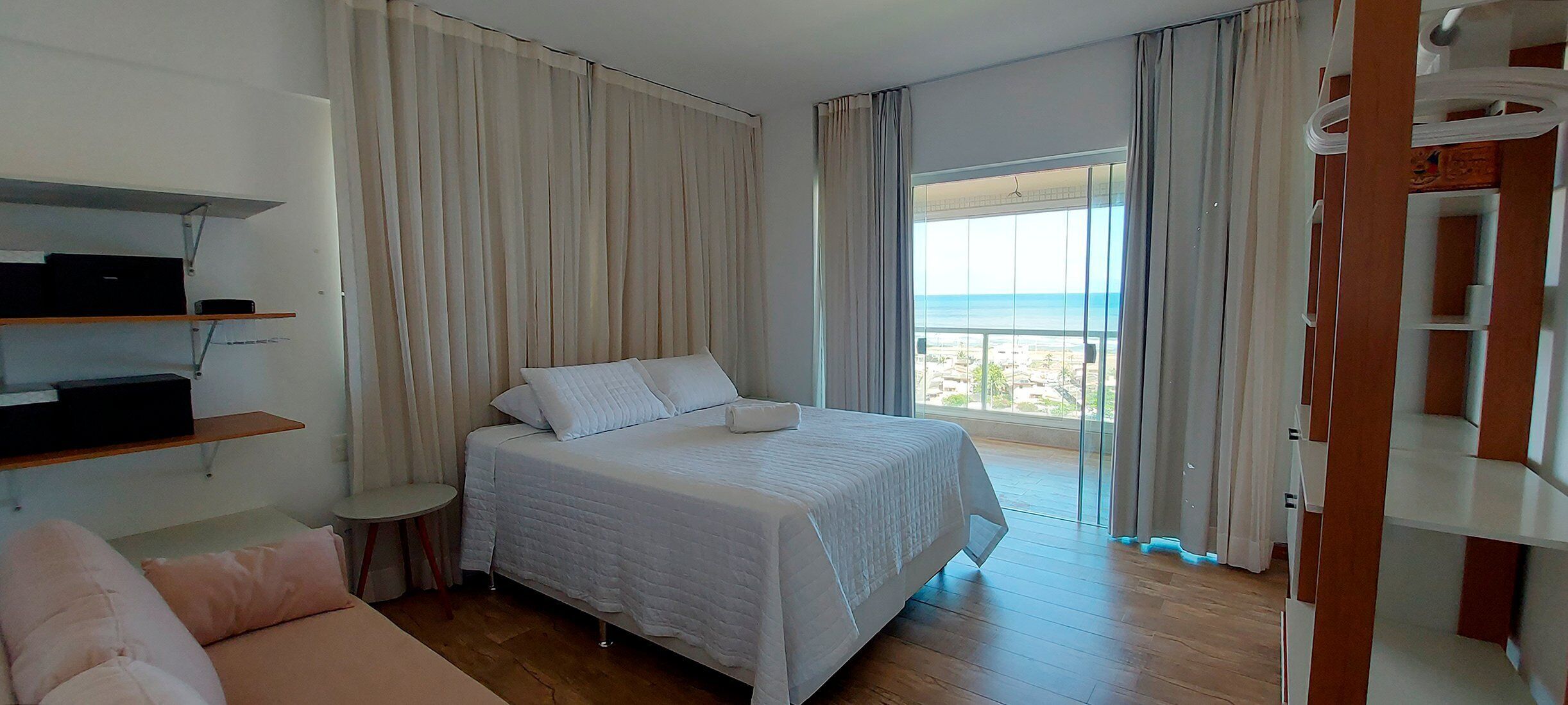Un dormitorio con cama, sofá, televisión en un estante y balcón con vista al mar.