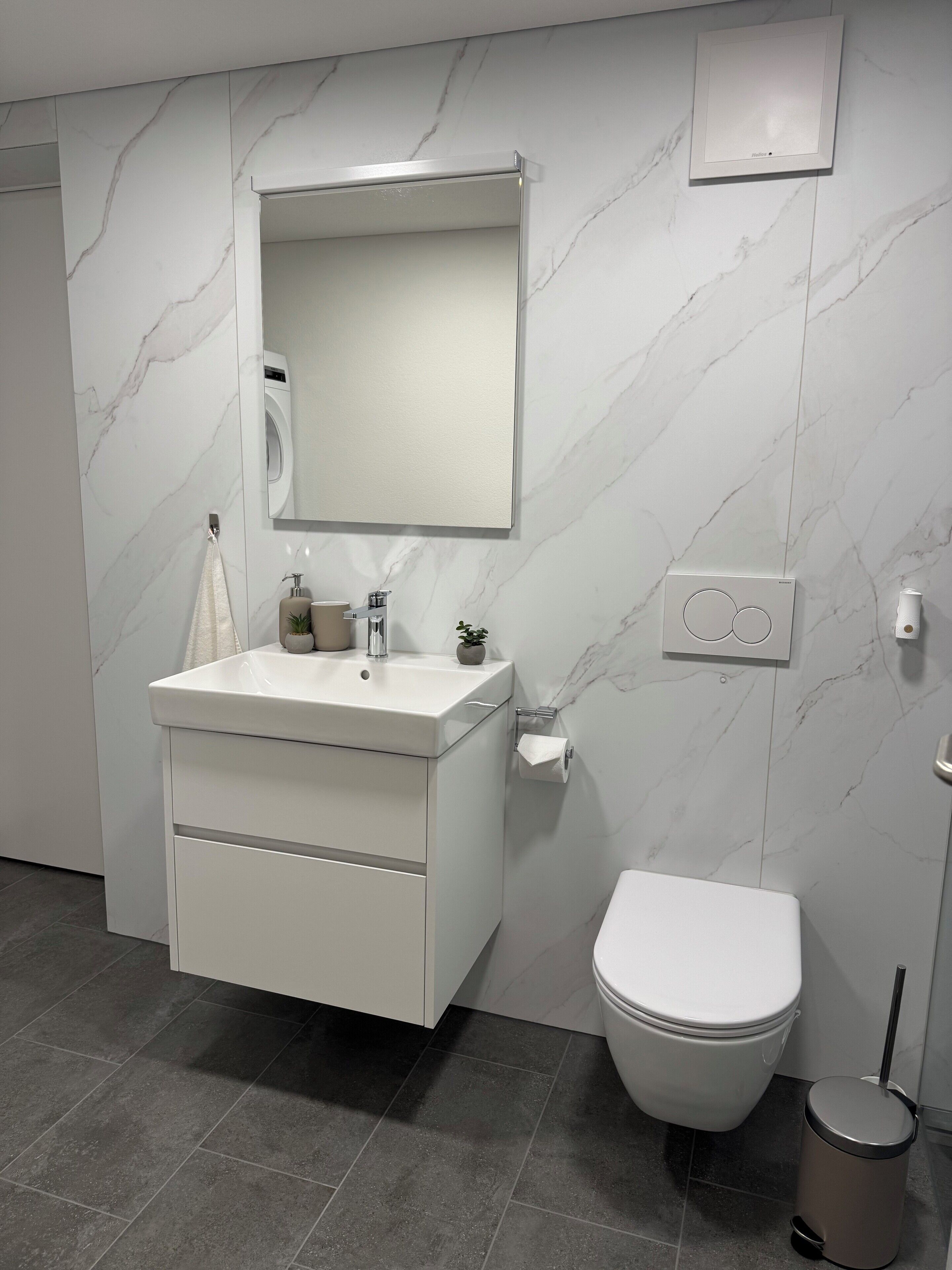 Een moderne badkamer met een wit badkamermeubel, een spiegel, een toilet en een prullenbak.