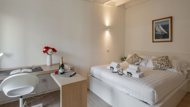 Una habitación de hotel moderna con escritorio, cama y toallas ordenadas, un cuadro en la pared y un jarrón con flores.