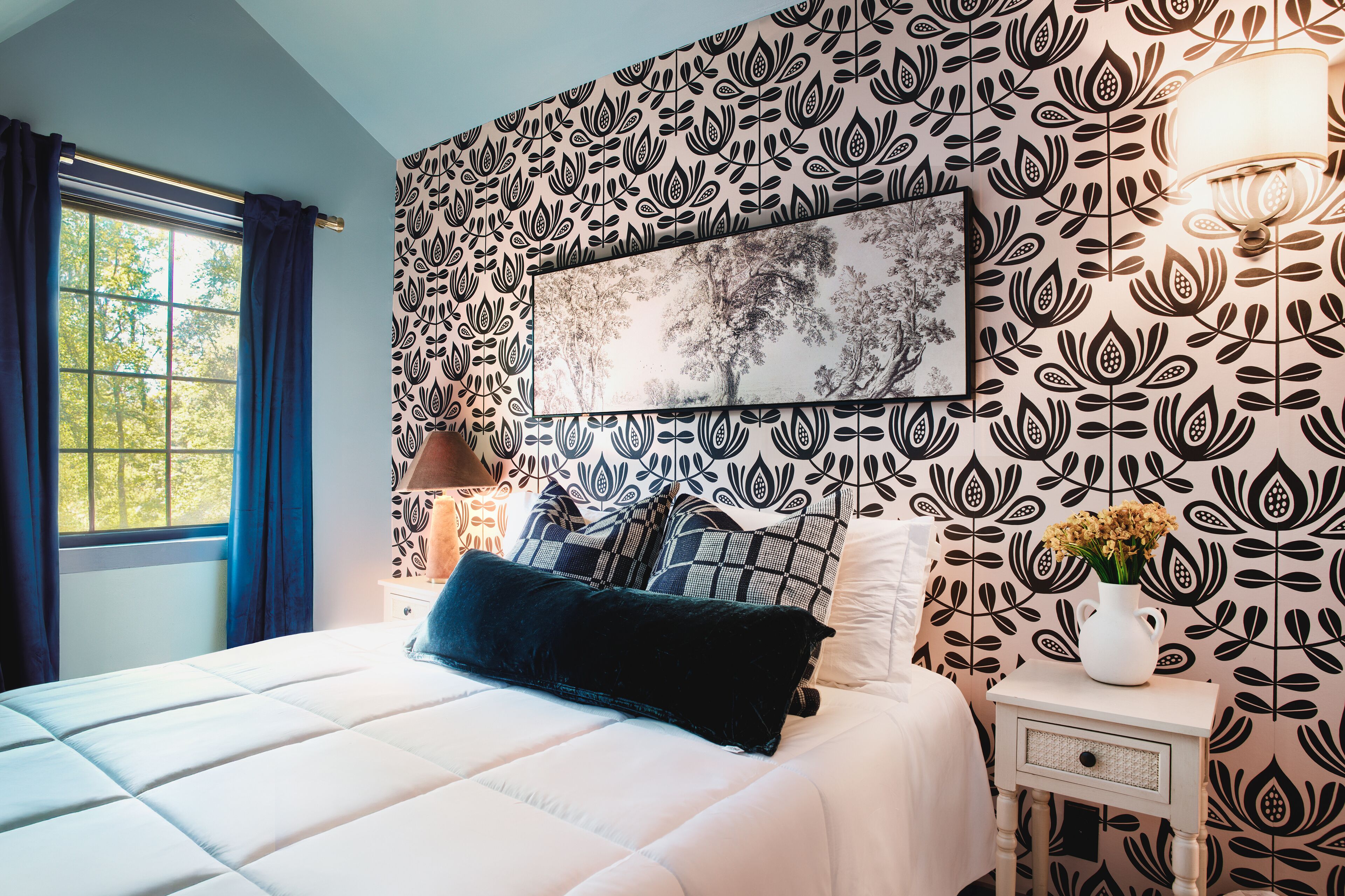 A bedroom with a patterned wallpaper, a bed with white bedding, a nightstand with a lamp, and a framed picture on the wall.