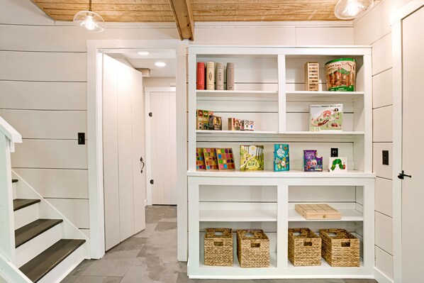 Un mueble blanco con estantes que contiene libros, juegos de mesa y cestas de almacenamiento en un pasillo.