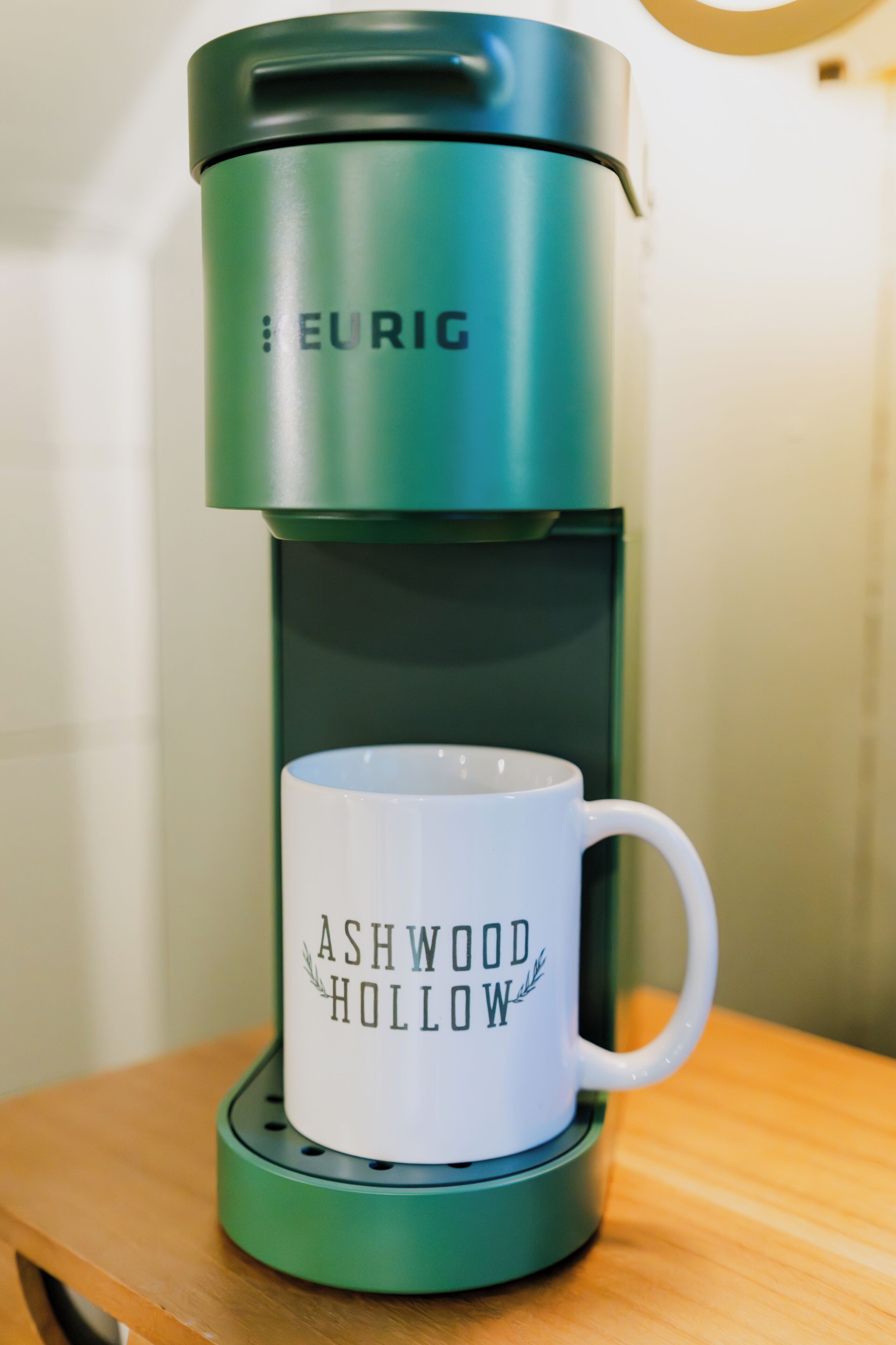 Una cafetera verde con una taza blanca que dice "Ashwood Hollow" sobre una mesa de madera.
