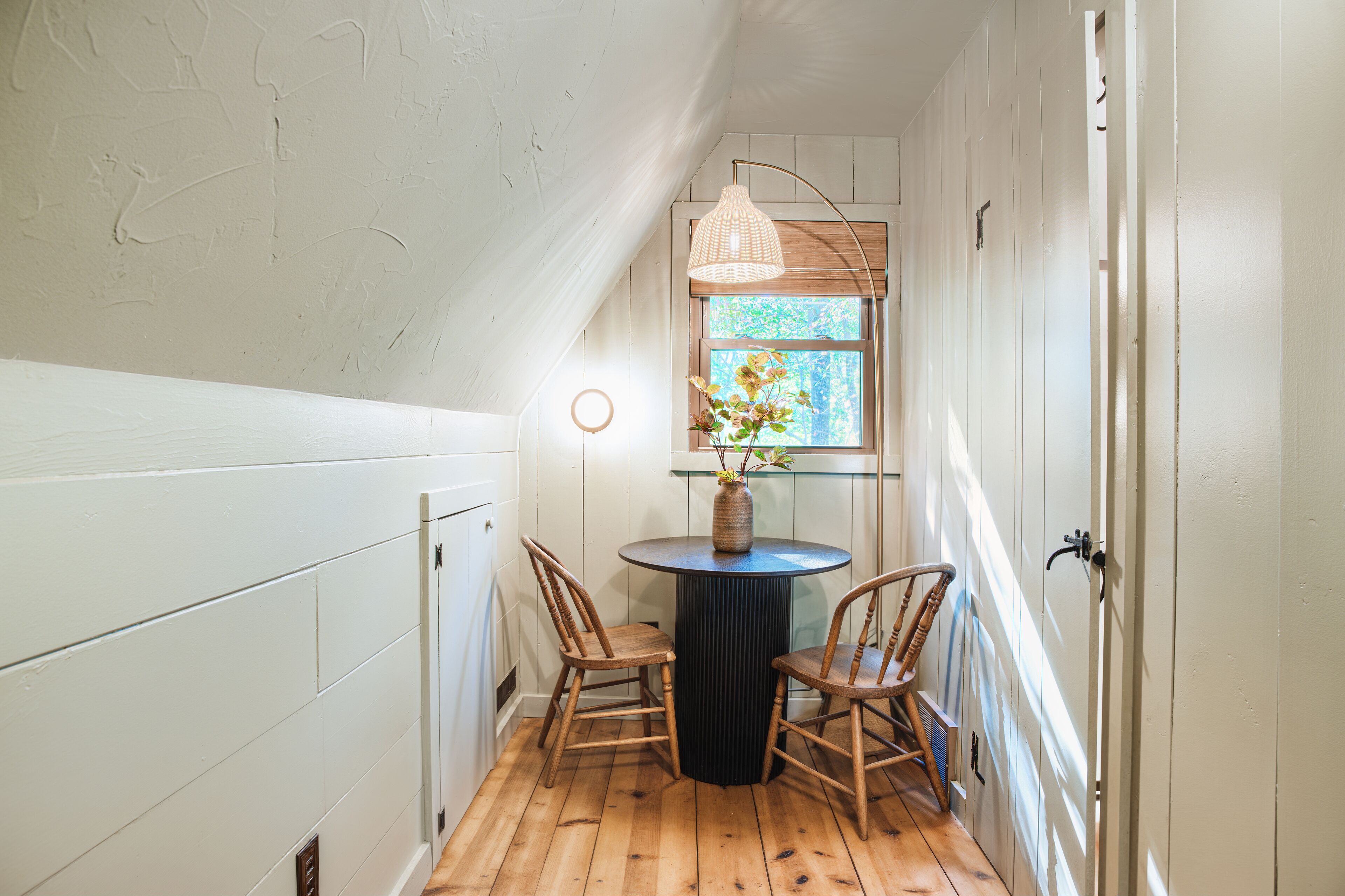 Un couloir étroit aux murs blancs, doté d’un plancher en bois et d’une table ronde accompagnée de deux chaises. On aperçoit un luminaire suspendu et une fenêtre donnant sur un paysage verdoyant.