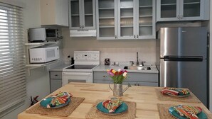A kitchen with white cabinets, a stainless steel refrigerator, a microwave, and a stove. There is a wooden table with colorful placemats and a vase with flowers. - Peaceful 2-bedroom condo in Wildwood Crest, close to beach (Wildwood)