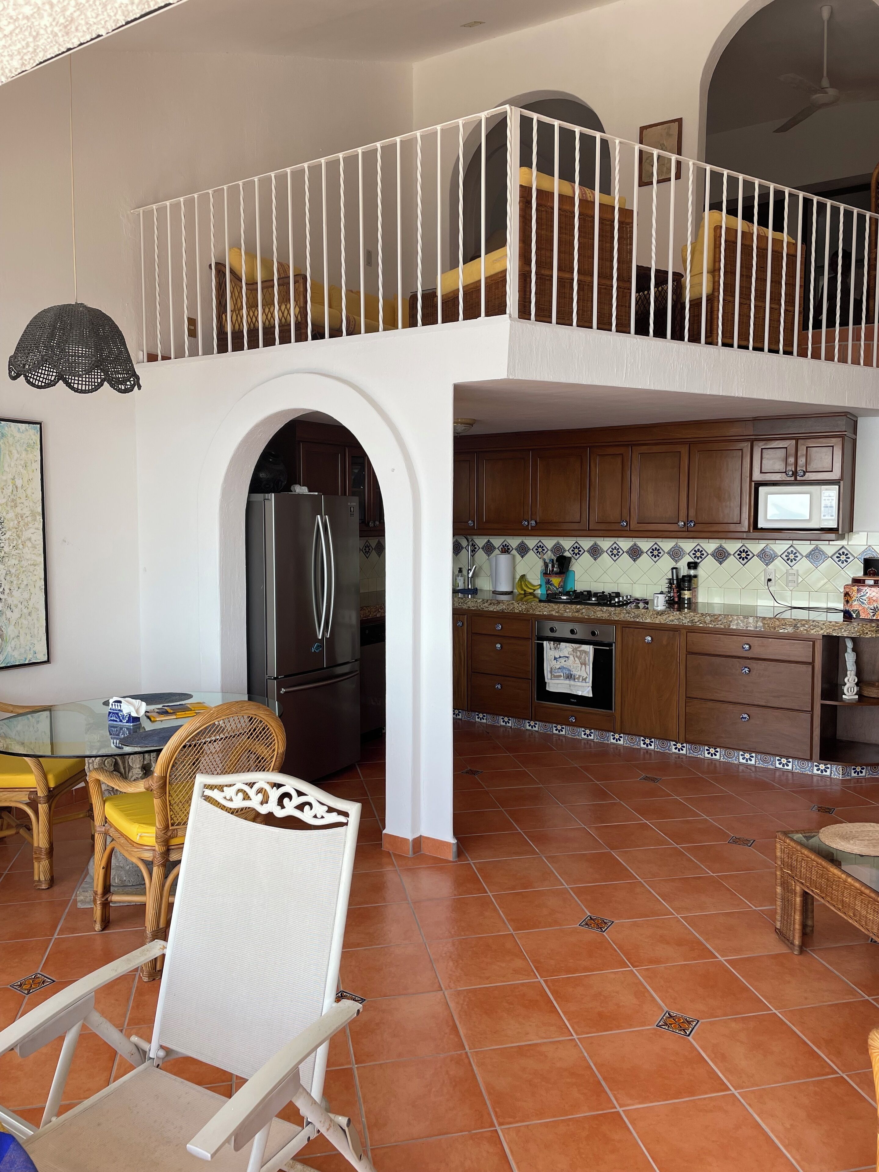 A spacious kitchen with wooden cabinets, a refrigerator, and a microwave. There is a dining area with a glass table and wicker chairs.