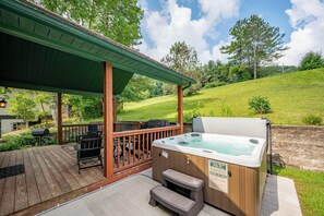 Un bain à remous sur une terrasse en bois, avec un espace couvert, du mobilier d’extérieur et une colline verdoyante en arrière-plan.