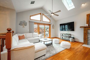 A modern living room with a white sofa, a coffee table, and a TV. There is a ceiling fan, a window, and a fireplace. - New! Modern Lakeview Ski Retreat w/ Hot Tub (McHenry)