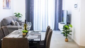 Ein Essbereich mit Tisch für vier Personen, eine Couch im Hintergrund und ein großes Fenster mit Vorhängen.