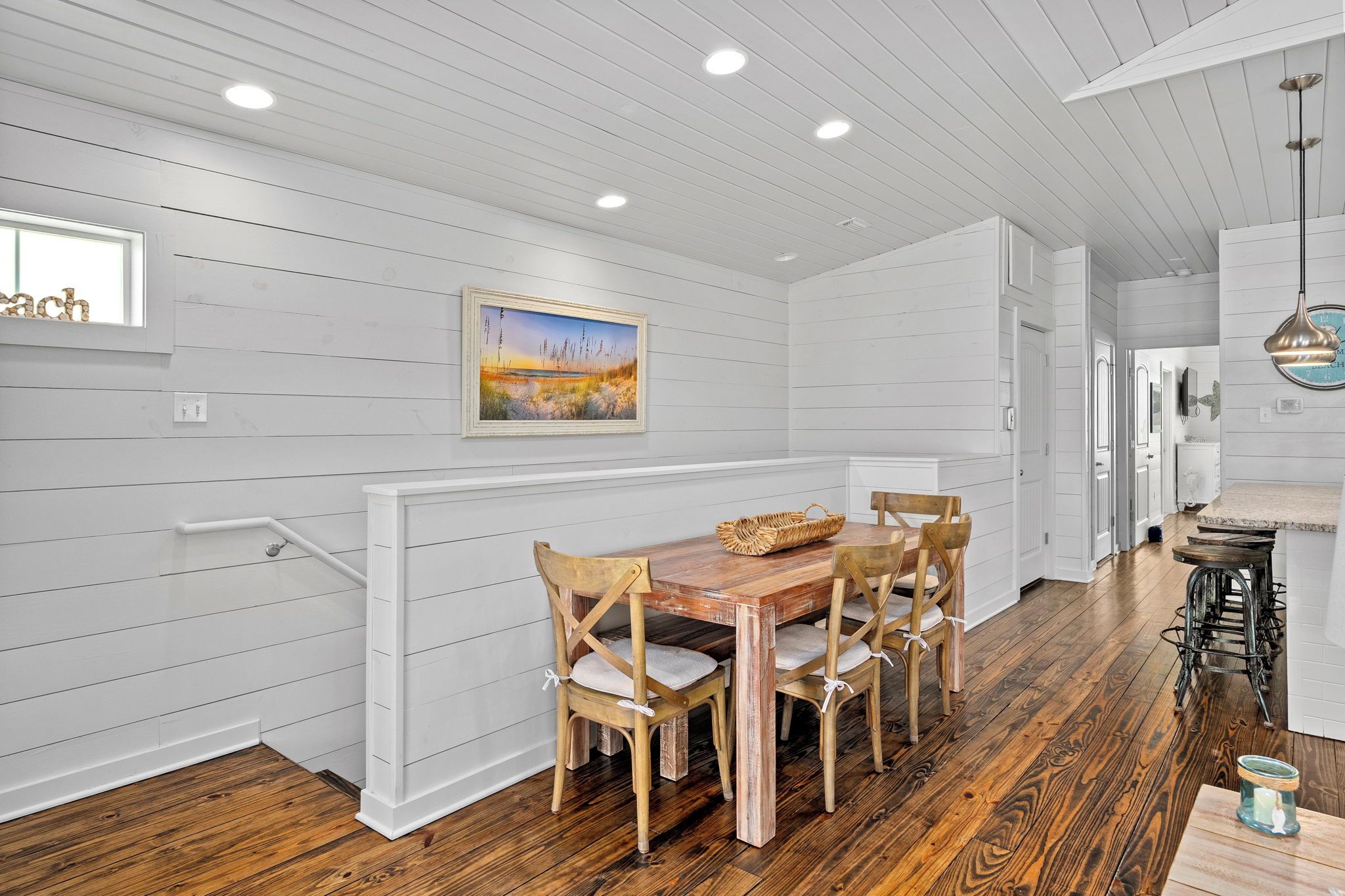 A dining area with a wooden table and chairs, white paneled walls, and a painting above the table.