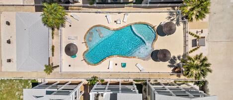An aerial view of a resort with a central swimming pool, multiple buildings, and palm trees.