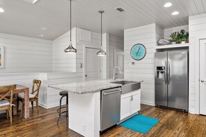 A kitchen with white cabinetry, a stainless steel refrigerator, pendant lights, and a clock on the wall. - Dauphin Beach House (Port Aransas)