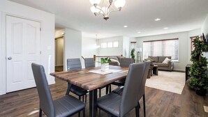 A modern living room with a dining area, featuring a wooden dining table, black chairs, a sofa, and a staircase leading to another level. - Chic 3BR Modern Home - Wi-Fi - Netflix - Sleeps 8! (Edmonton)