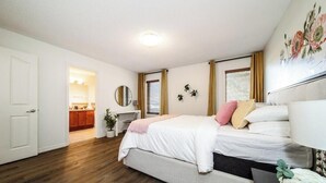 A bedroom with a bed, bedside table, and a mirror. There is a door leading to a bathroom and a kitchen area. - Chic 3BR Modern Home - Wi-Fi - Netflix - Sleeps 8! (Edmonton)