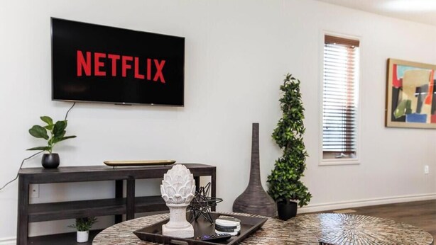 A living room with a mounted TV displaying the Netflix logo, a dark wooden console table, a round wicker coffee table, and a tall vase. - Chic 3BR Modern Home - Wi-Fi - Netflix - Sleeps 8! (Edmonton)