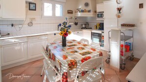 Eine Küche mit weißen Schränken, einem Esstisch mit einer Blumen Tischdecke und einer Kücheninsel mit einem Rollwagen.