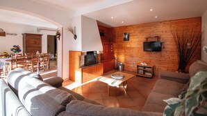 Ein Wohnzimmer mit einer Couch, einem Couchtisch und einem Fernseher. Im Hintergrund ist eine Holzwand und ein Essbereich zu sehen.