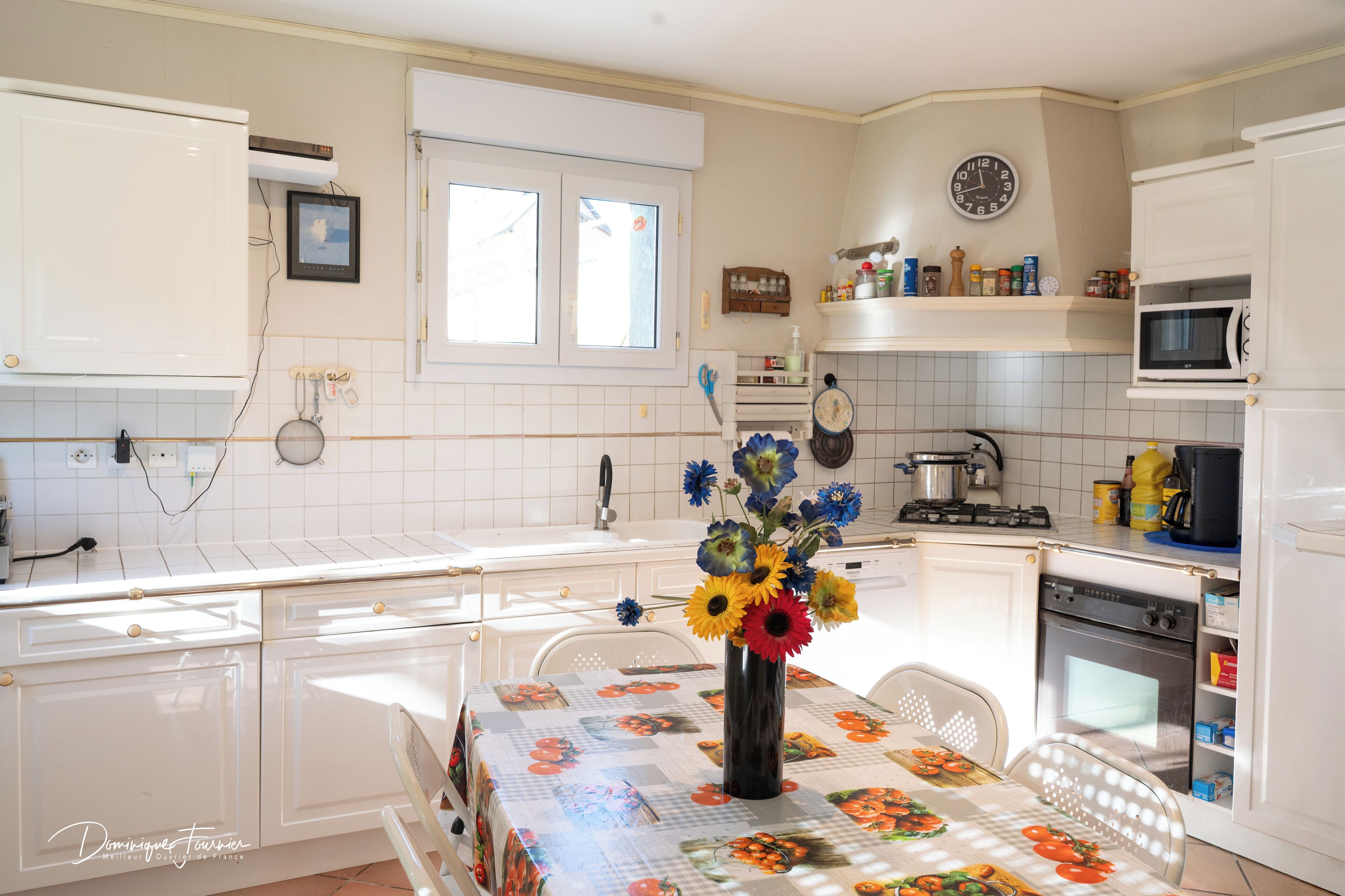 A kitchen with white cabinets, a dining table with a floral tablecloth, and a vase with flowers.