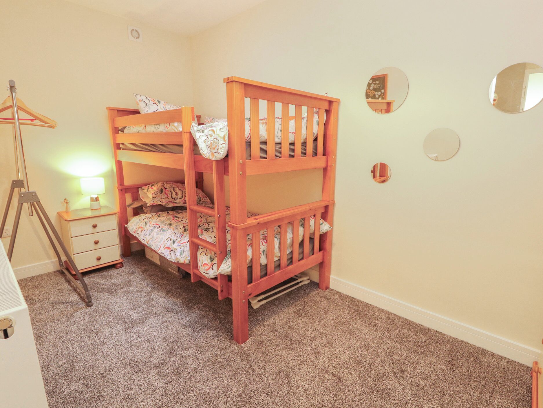 A bunk bed with a small bedside table and lamp, a tripod lamp, and a mirror on the wall.
