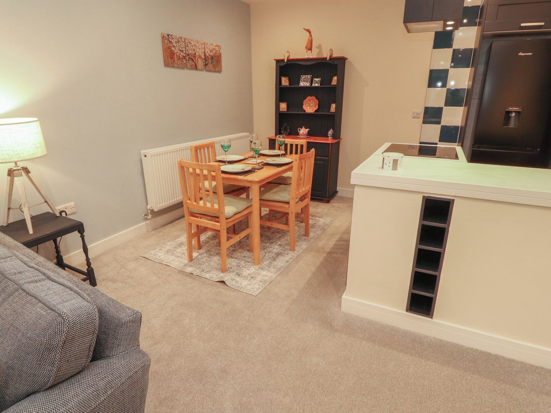 A compact kitchen with a dining table, chairs, a radiator, a refrigerator, and a small cabinet with decorative items.