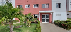 A building with a red facade, a sign reading "PúPura," and a well-maintained garden with palm trees and flowers. - COPIA0102 - Cond Cores de Piatã (Salvador)
