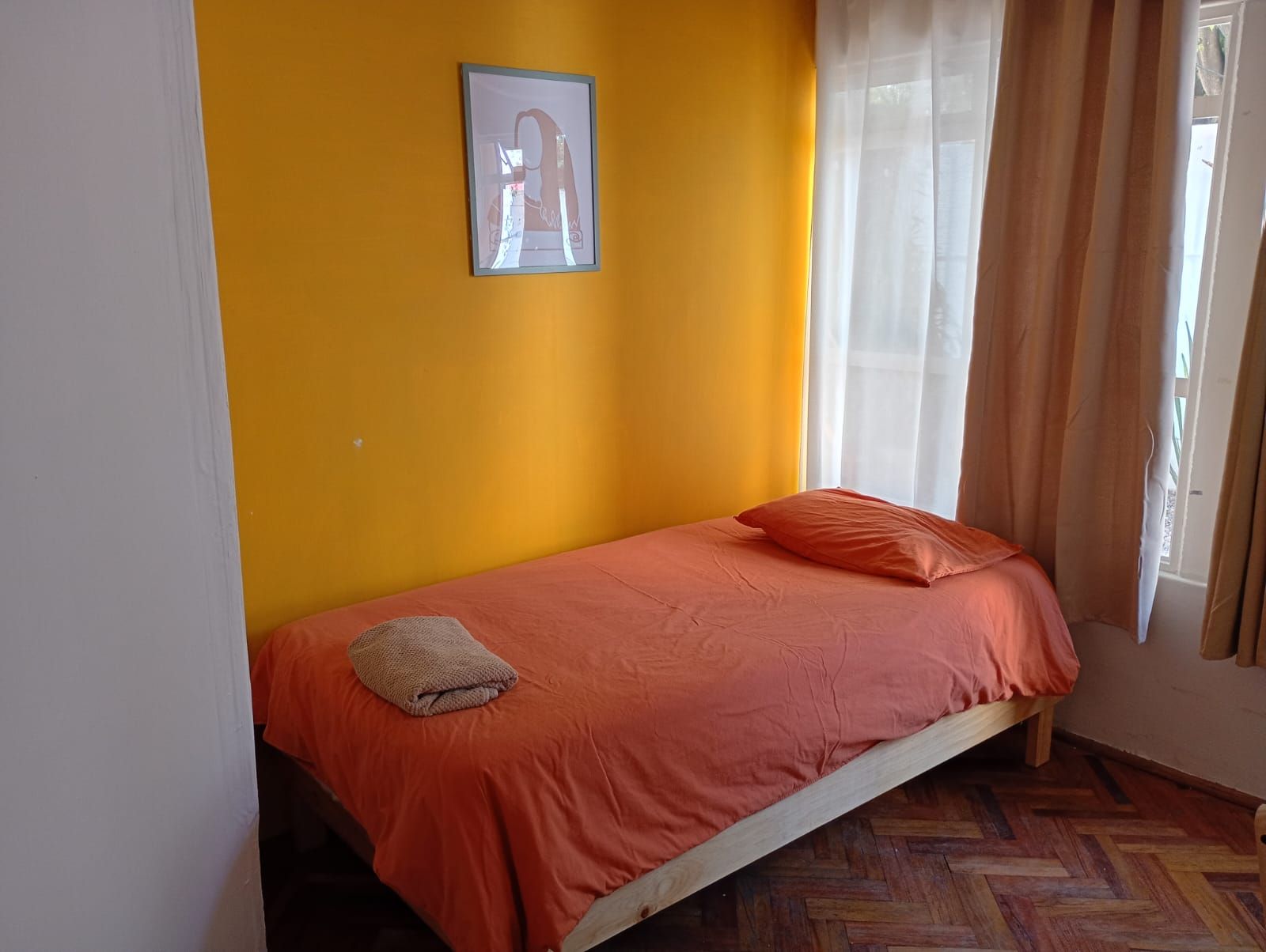 Una cama individual con colcha y almohadas naranja, un cuadro enmarcado en la pared y una ventana con cortinas.