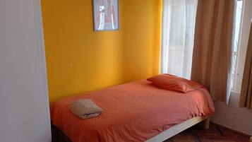 Una cama individual con colcha y almohadas naranja, un cuadro enmarcado en la pared y una ventana con cortinas.