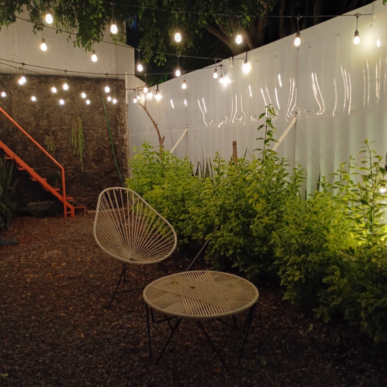 Un área de estar al aire libre con dos sillas y una mesa, rodeada de follaje y una pared blanca adornada con luces colgantes.