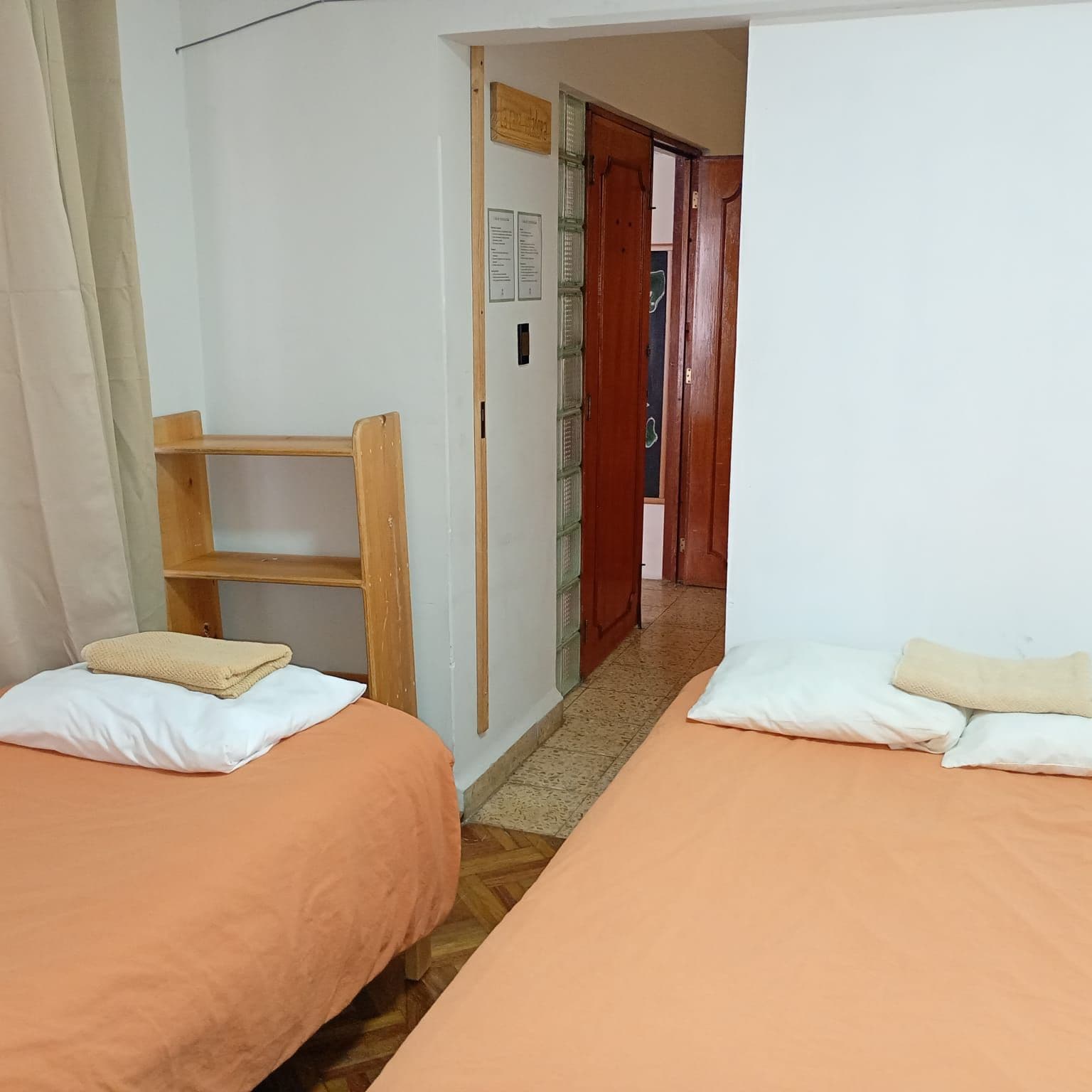 Una habitación con dos camas, cada una con una almohada y una manta, una silla de madera y una puerta que da a otra habitación.