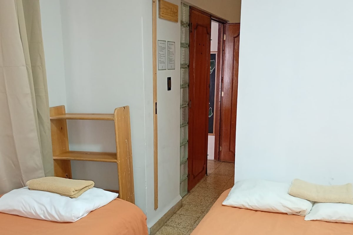 A room with two beds, each with a pillow and a blanket, a wooden chair, and a door leading to another room.