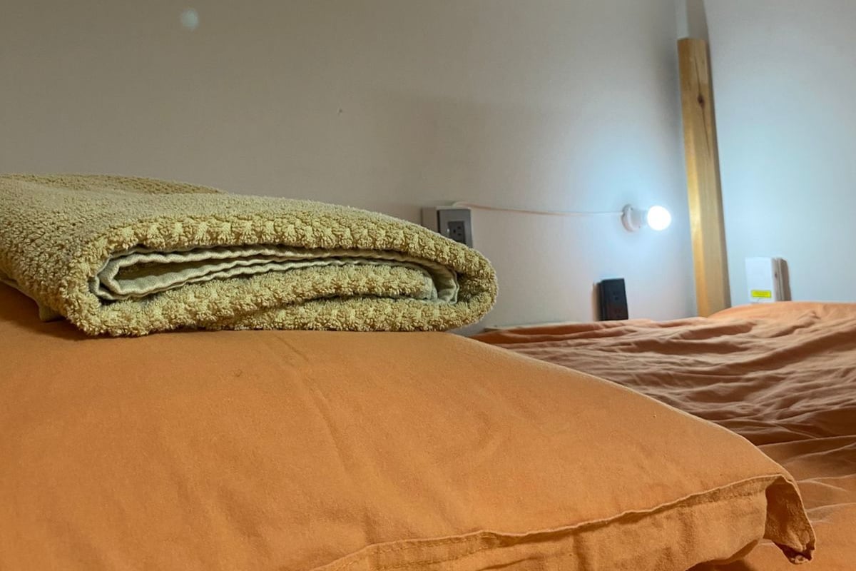 A hotel room with a bed, a folded blanket, a bedside table with a lamp, and a wall-mounted light.