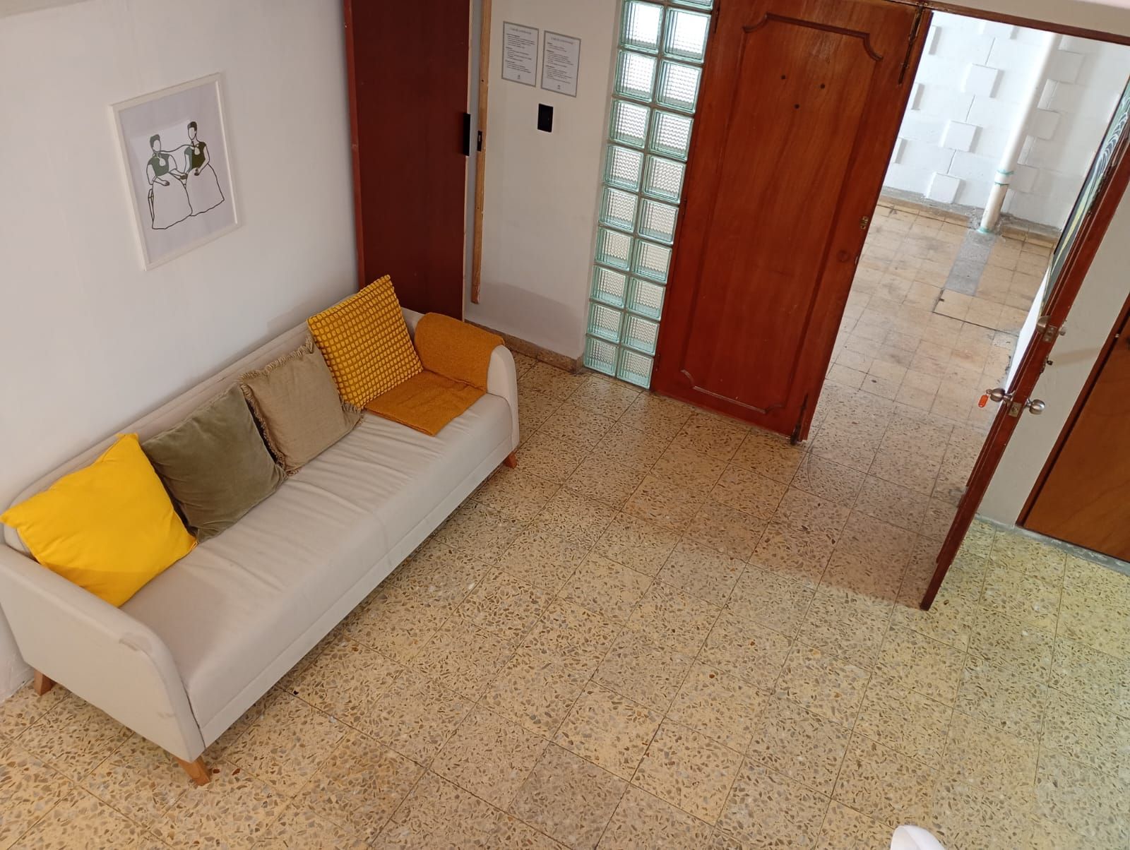 Una habitación con un sofá blanco, una puerta de madera y piso de baldosas.