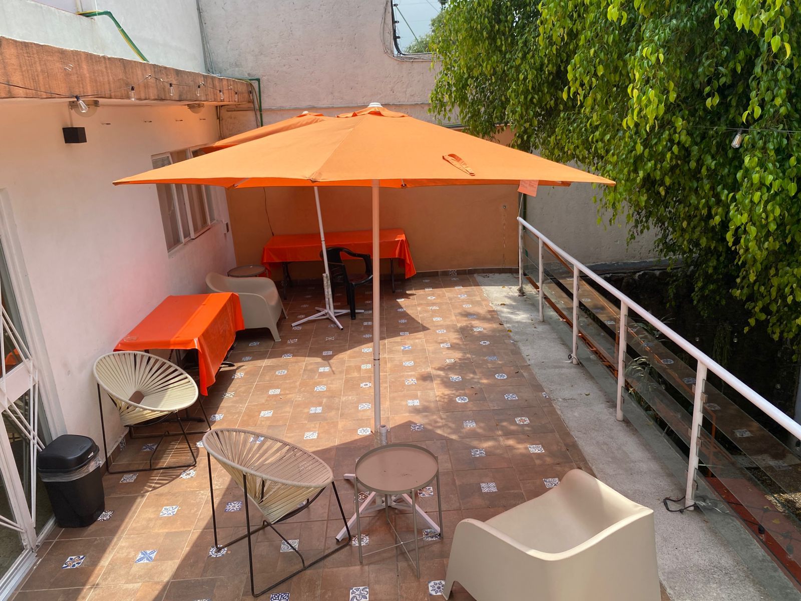 Eine Terrasse im Freien mit Fliesenboden, einem Tisch mit orangefarbener Tischdecke, zwei Korbstühlen und einem großen orangefarbenen Sonnenschirm.