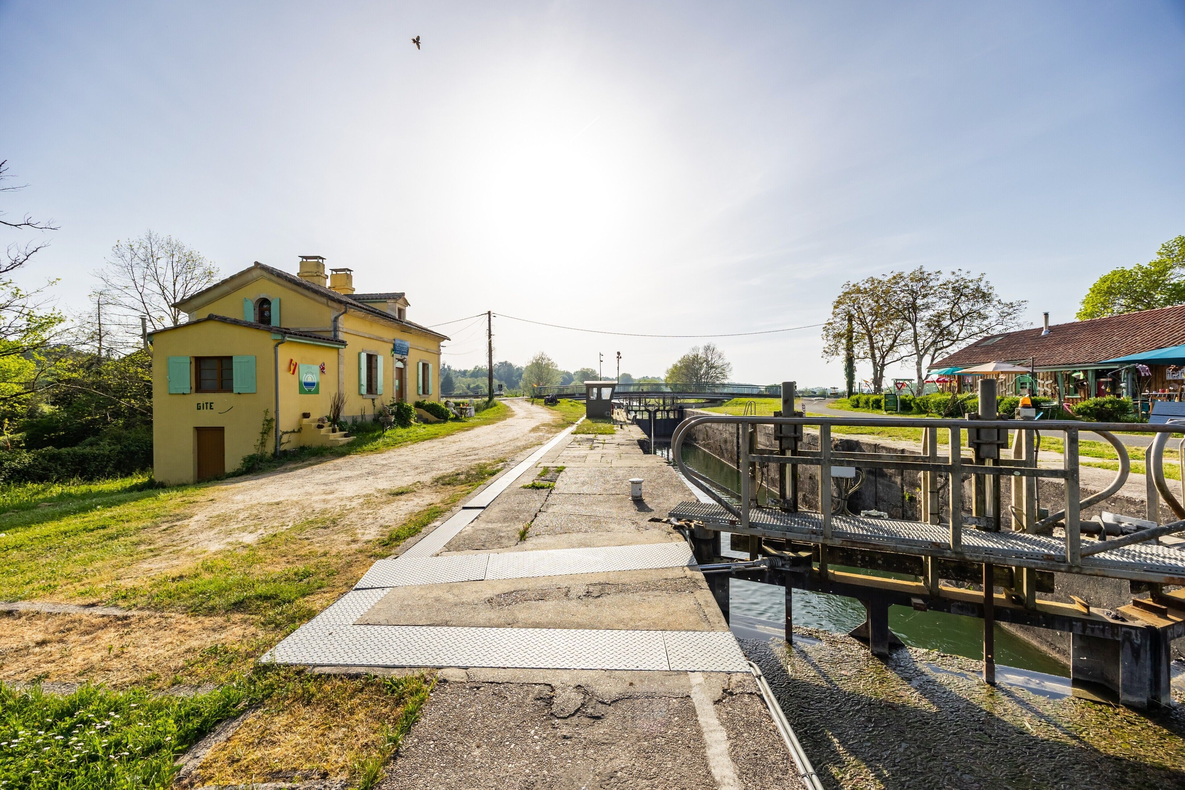 Un canal surmonté d’un pont piétonnier, un bâtiment jaune portant une enseigne « GÎTE », et une petite construction avec un toit bleu.