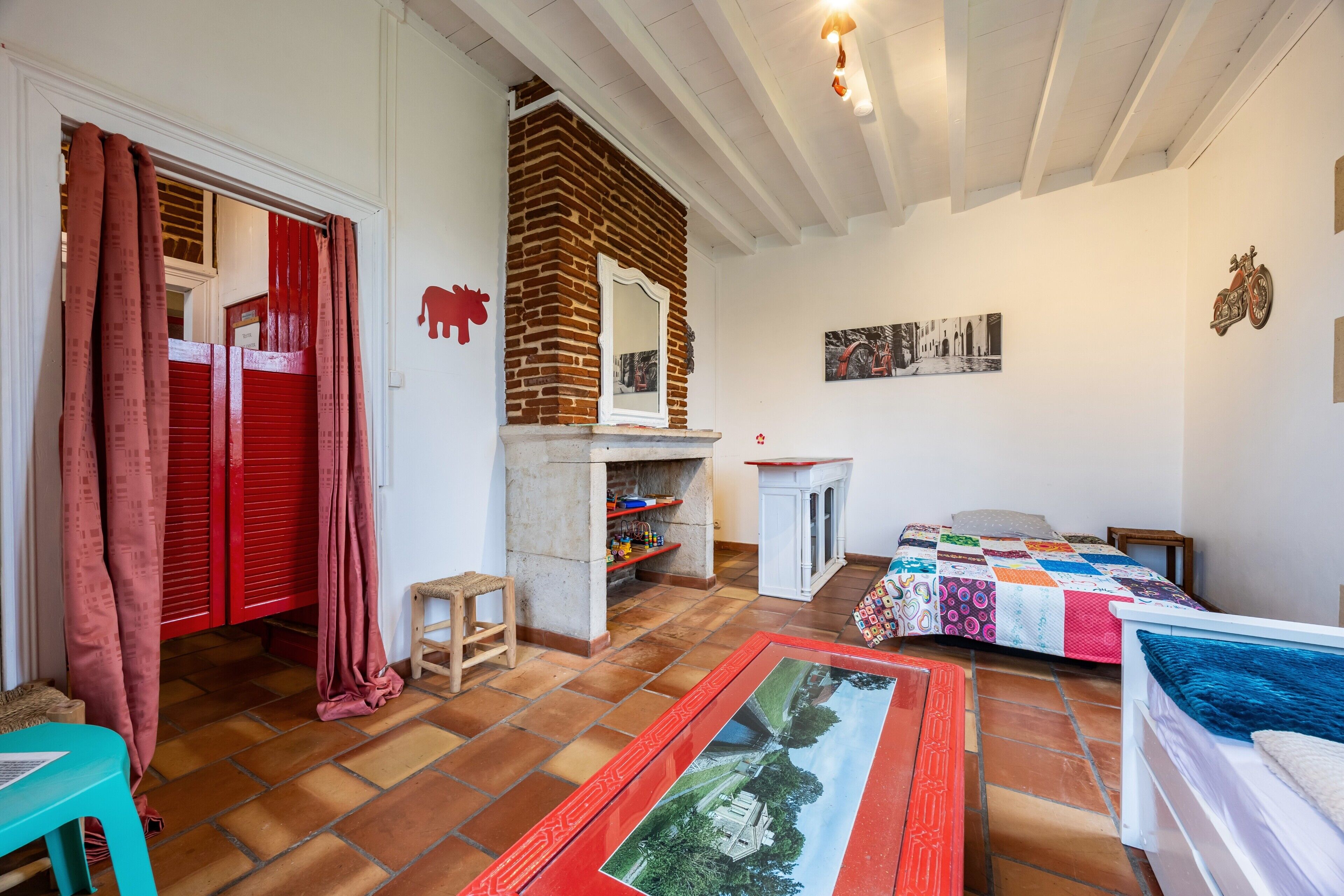 Une pièce avec une cheminée, un lit recouvert d’une couverture aux couleurs vives, une porte rouge et un sol carrelé.