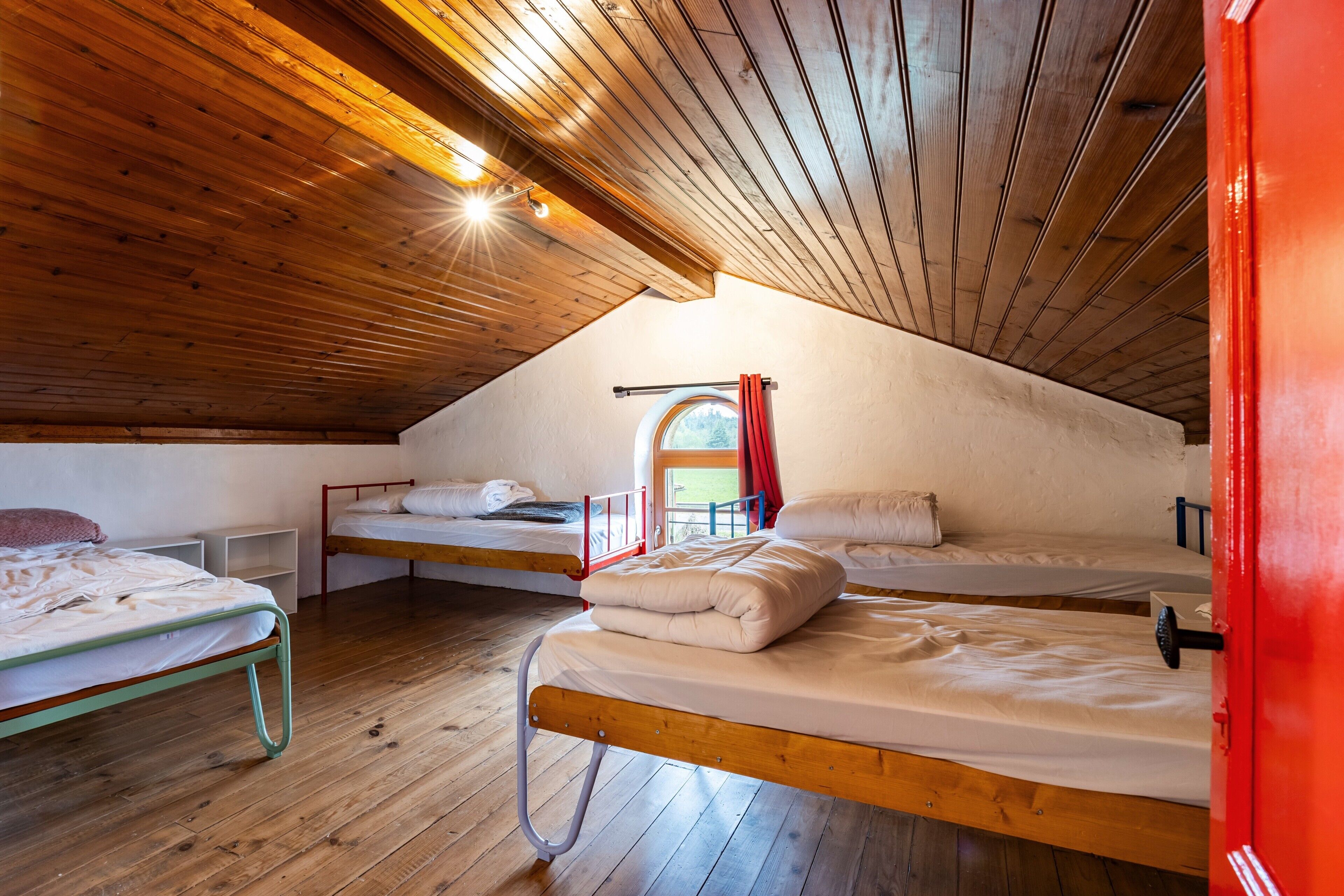 A dormitory room with wooden ceilings, bunk beds, a window with a view, and a red door.