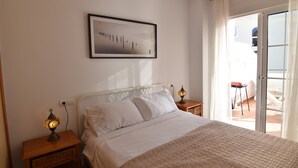 A bedroom with a bed, two bedside tables with lamps, a framed picture on the wall, and a sliding glass door leading to an outdoor area. - Corinto Apartment, 100m from Burriana Beach (Nerja)