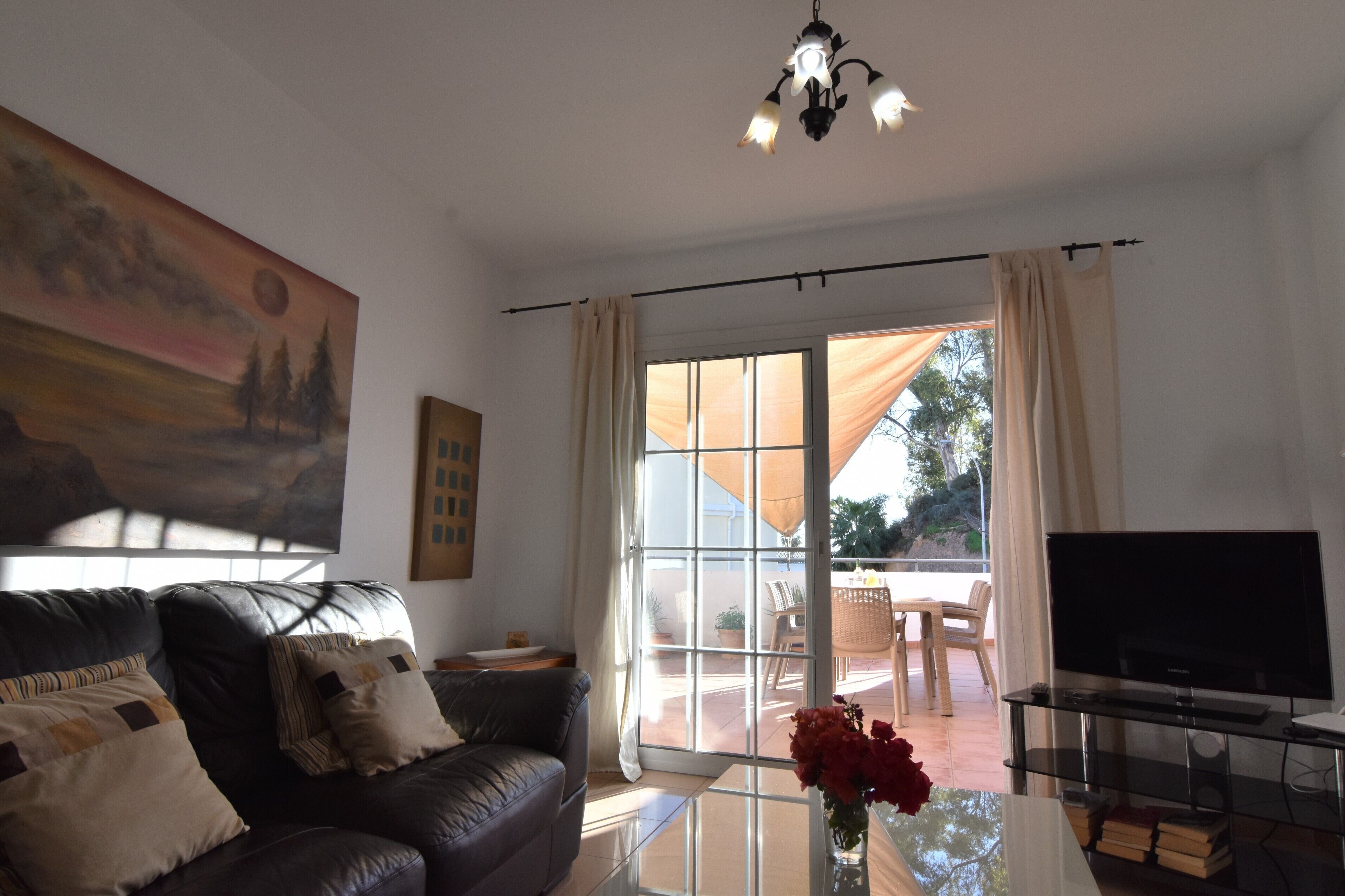 A living room with a leather sofa, a glass coffee table with books and flowers, a flat-screen TV, and a large painting on the wall.