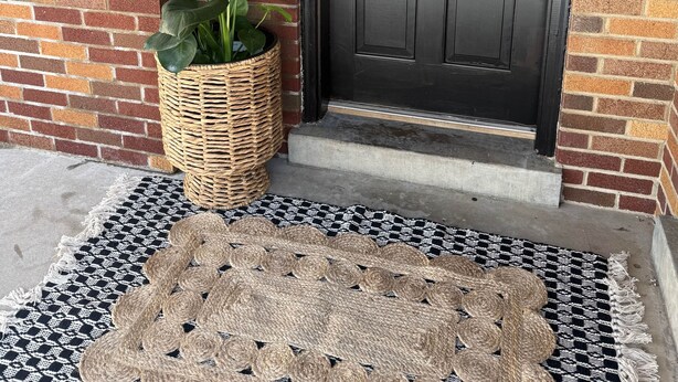 Tumma puuovi, jossa on koristeellinen seppele, ruukkuistutettu kasvi ja kudottu ovimatto.
