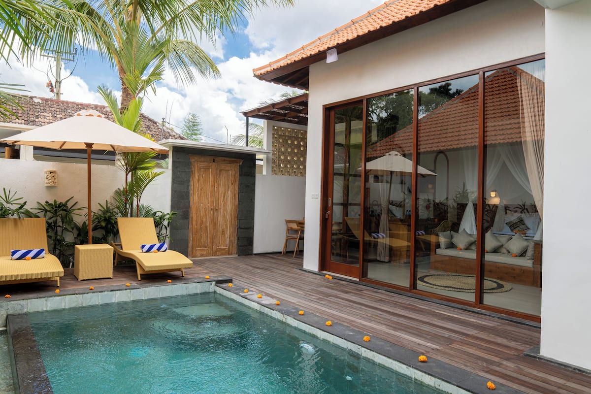 Un espace piscine comprenant des chaises longues, une terrasse en bois et une pièce fermée par des parois de verre, avec un canapé et des rideaux.