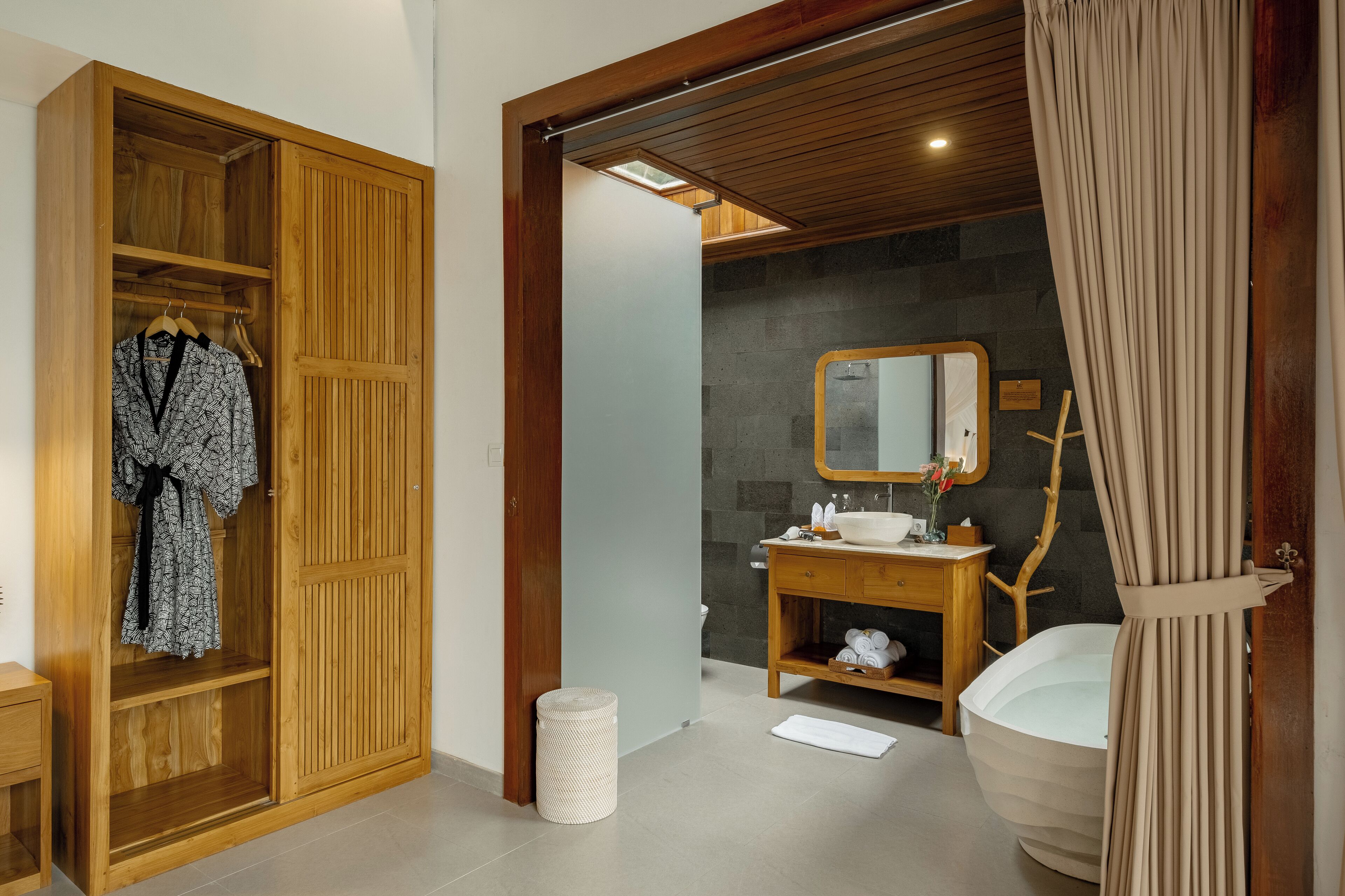 A bathroom with a wooden vanity, a large mirror, a bathtub, and a wooden wardrobe with a robe hanging.