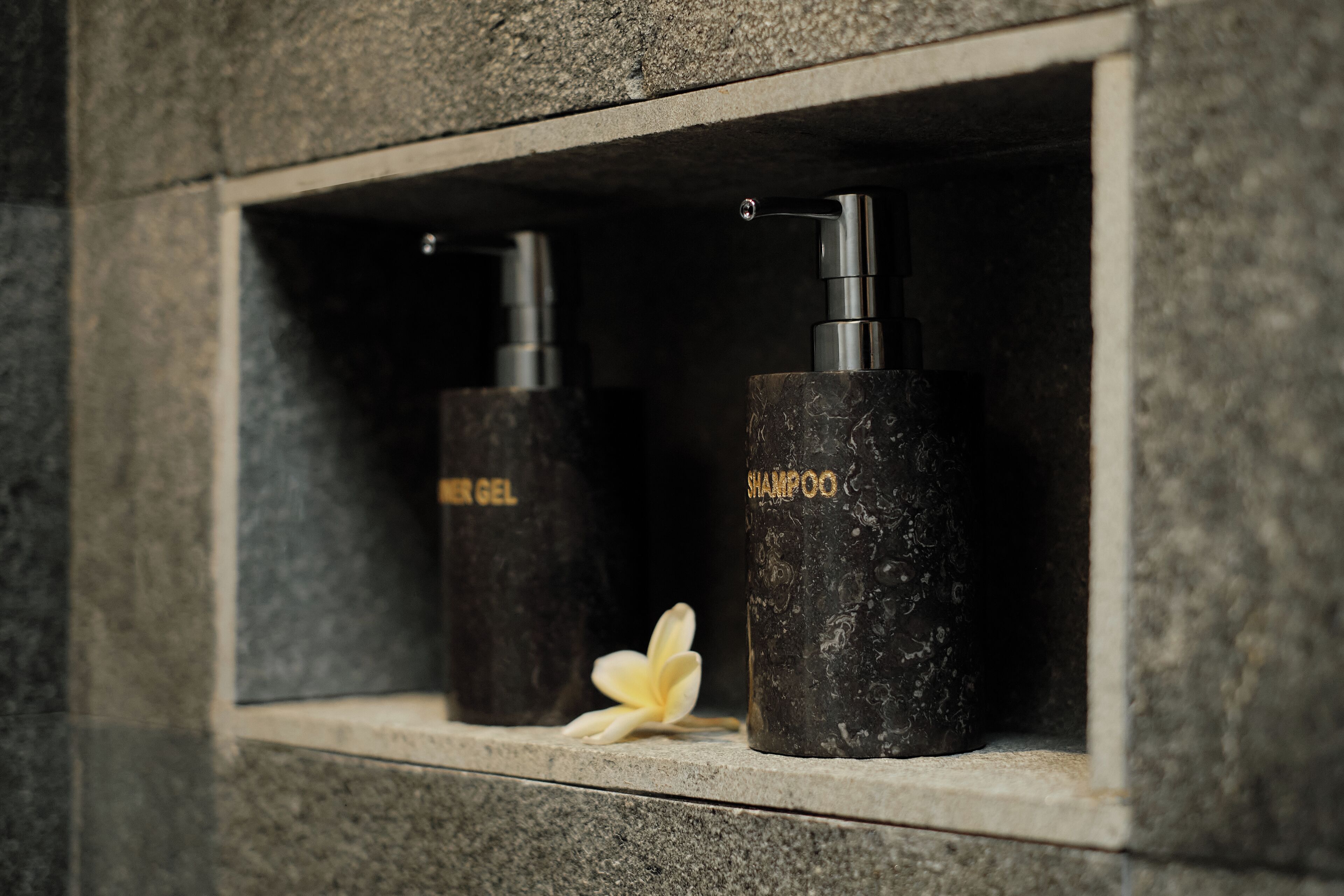 Une étagère de salle de bain avec deux bouteilles de shampoing et d’après-shampoing, et une fleur blanche.