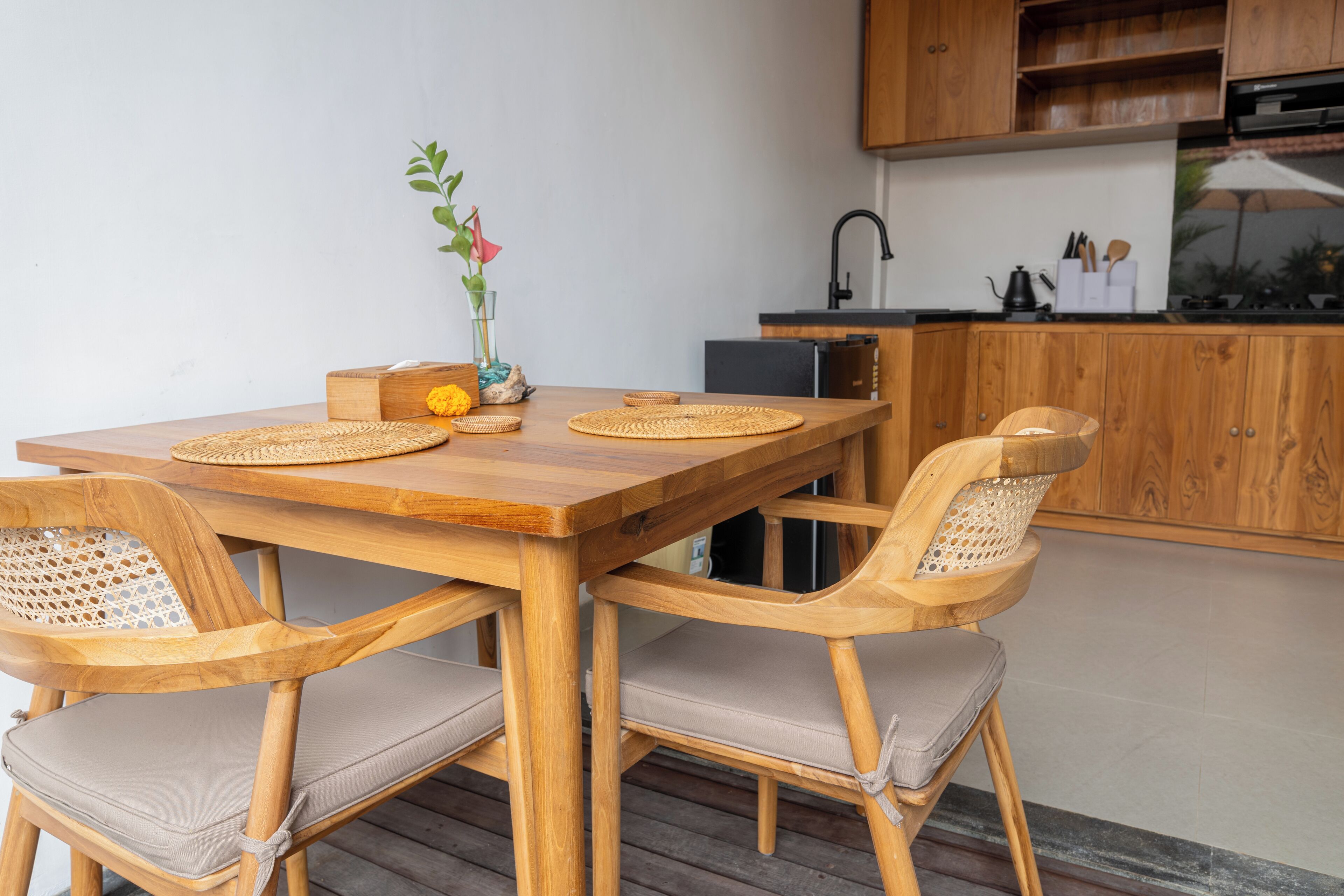 Una mesa de comedor de madera con individuales tejidos, dos sillas acolchadas y un jarrón con flores.