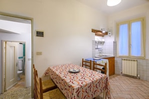 Una cocina compacta con una mesa de comedor, sillas, una ventana con cortinas, un radiador y la puerta del baño a la vista.