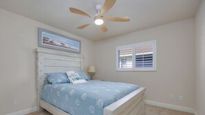 Un dormitorio con cama, ventilador de techo, ventana con persianas e imagen colgada en la pared.