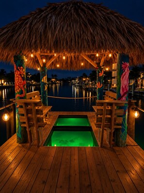 A wooden deck with a thatched roof, surrounded by water, and illuminated by string lights. - Gulf Access•Glass-Bottom Tiki•FREE Heated Pool•Smart TVs•Kayaks•Beach/Fish Gear (Cape Coral)