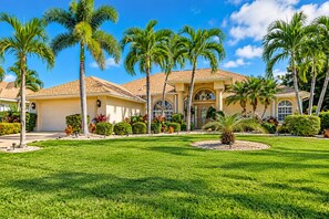 A residential house with a well-maintained lawn, palm trees, and a garage. - Gulf Access•Glass-Bottom Tiki•FREE Heated Pool•Smart TVs•Kayaks•Beach/Fish Gear (Cape Coral)